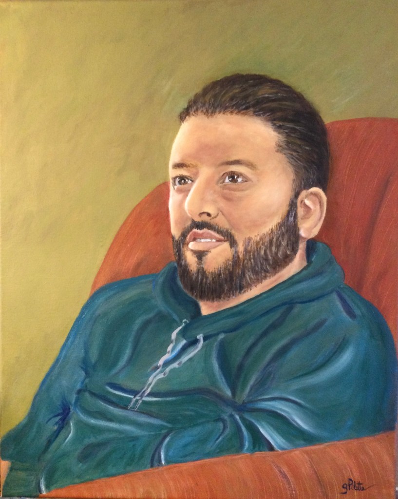 Jeune homme barbu aux cheveux et yeux bruns, vêtu de bleu dans un fauteuil orangé sur un fond jaunâtre 